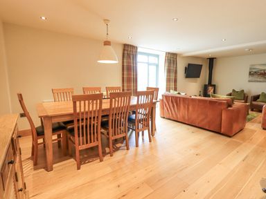 A dining area with wooden table and chairs next to a living room with leather sofas a wood burning stove and a wall mounted tv at Yew Cottage in Backbarrow