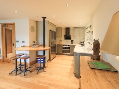 A kitchen with wooden floor stools a circular wooden table stainless steel fridge and oven at Yew Cottage Backbarrow