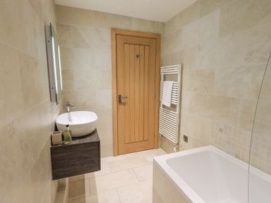 A bathroom with a sink on a floating wooden vanity a bathtub a wooden door and a towel radiator with a towel at Yew Cottage in Backbarrow