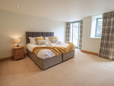 A bedroom with a double bed with pillows and a blanket bedside tables with lamps and windows with curtains at Yew Cottage in Backbarrow