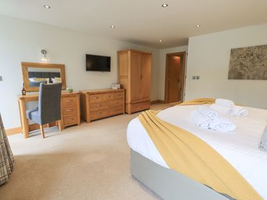 A bedroom with a bed with towels and a yellow throw a desk with a grey chair a dresser a wardrobe and a wall mounted tv at Yew Cottage in Backbarrow