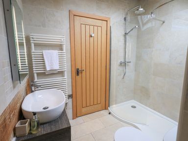 A bathroom with a wooden door a white vessel sink on a dark wooden cabinet a white towel on a wall-mounted radiator and a corner shower with glass partition at Yew Cottage in Backbarrow
