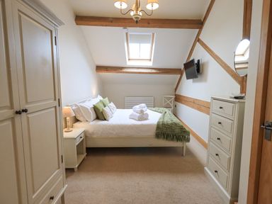 A bedroom with a bed with pillows and folded towels a wardrobe a chest of drawers a chair and a wall mounted tv at Yew Cottage in Backbarrow