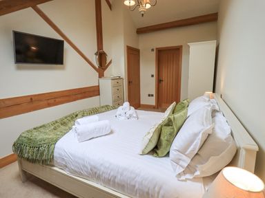 A bedroom with a neatly made bed with green pillows and folded towels a wall-mounted tv and wooden doors at Yew Cottage in Backbarrow