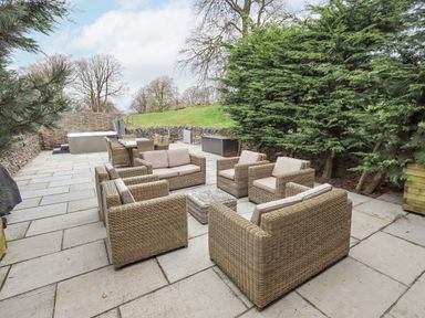 An outdoor patio area with wicker chairs and sofas around a glass table a dining table with chairs and a hot tub surrounded by trees and stone walls at Yew Cottage in Backbarrow