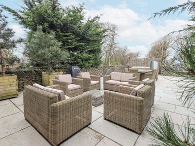 An outdoor patio with wicker armchairs sofas a small glass table large planters with trees and a dining table in the background at Yew Cottage in Backbarrow