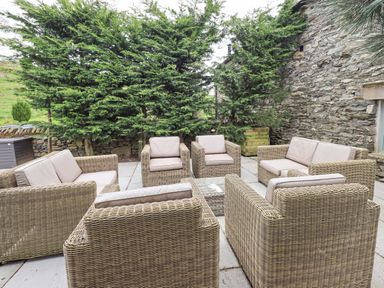 An outdoor patio area with six wicker chairs and two two-seater wicker sofas arranged around a small table with trees and a stone wall in the background at Yew Cottage in Backbarrow