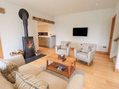 A living room with a wood stove two armchairs a sofa a wooden coffee table and a wall-mounted television at Lime Cottage in Backbarrow near Newby Bridge