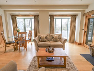 A living room with a sofa two windows wooden dining table and chairs at Lime Cottage in Backbarrow near Newby Bridge