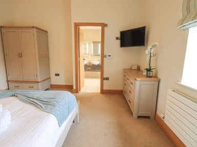 A bedroom with a bed, wardrobe, dresser with a plant, wall-mounted TV, and an open door to a bathroom at Lime Cottage in Backbarrow near Newby Bridge