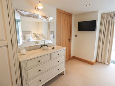 A bedroom corner with a white dresser a mirror a small plant a jar a wall-mounted television a wooden door and curtains at Lime Cottage in Backbarrow near Newby Bridge