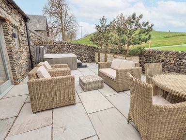 An outdoor patio with rattan furniture including chairs a sofa and a table a hot tub and planters with trees at Lime Cottage in Backbarrow near Newby Bridge