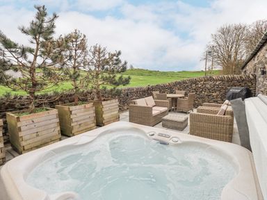 An outdoor patio with a hot tub wooden planters wicker seating and a stone wall at Lime Cottage in Backbarrow near Newby Bridge
