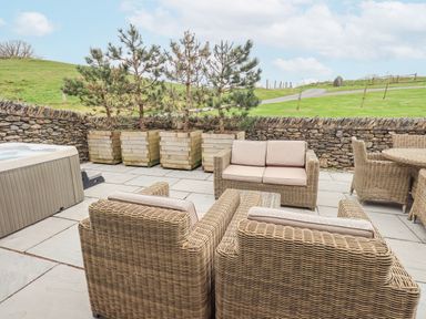 An outdoor patio with wicker chairs and sofa a hot tub and wooden planters with trees at Lime Cottage in Backbarrow near Newby Bridge