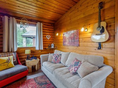 A living room with a couch pillows a guitar on the wall a window with curtains and a small side table at Harbottle Lodge in Longframlington near Alnwick