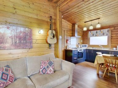 A wooden living room and kitchen with a sofa with patterned cushions a guitar on the wall and a dining table with chairs at Harbottle Lodge in Longframlington near Alnwick