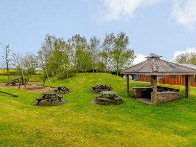 An outdoor picnic area with wooden tables benches a covered brick barbecue and a children's play area at Harbottle Lodge in Longframlington near Alnwick