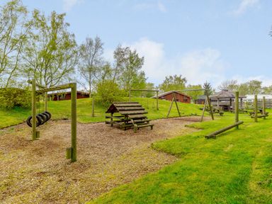 An outdoor playground with wooden climbing structures swings and picnic tables surrounded by grass and trees at Harbottle Lodge in Longframlington near Alnwick