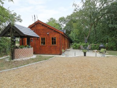 An outdoor area with a wooden cabin surrounded by trees a gravel ground and a patio with a table and chairs at Acorn Lodge in Saxmundham
