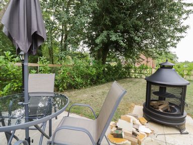 A garden patio with a glass table and chairs an umbrella a fire pit with firewood and trees at Acorn Lodge in Saxmundham