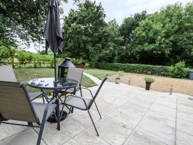 A patio with a round glass table and four chairs and a closed umbrella near a grass yard with trees and bushes at Acorn Lodge in Saxmundham