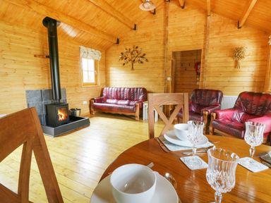 A wooden living room with red leather sofas a wood burning stove and a dining table with glassware at Acorn Lodge in Saxmundham