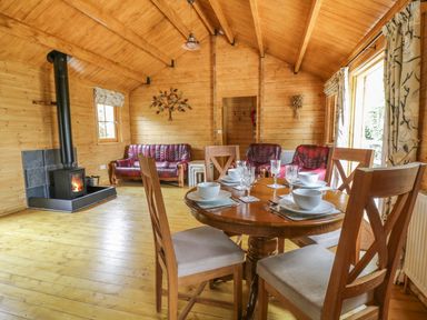 A wooden dining table set with bowls and glasses next to a wood burning stove and red leather sofas in a wooden cabin at Acorn Lodge in Saxmundham