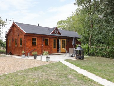 A wooden lodge with multiple windows and doors opening to a patio with a table, chairs, and an outdoor fireplace surrounded by grass and trees at Acorn Lodge in Saxmundham