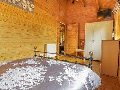 A bedroom with wooden walls a bed with floral bedding a wardrobe a chair a dresser and a wall-mounted television at Acorn Lodge in Saxmundham