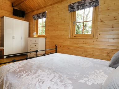 A bedroom with a bed a white wardrobe a chest of drawers two windows and a mounted television at Acorn Lodge in Saxmundham