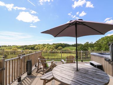 An outdoor deck with wooden chairs a round table with umbrella and a hot tub overlooking green hills at Foxes Meadow in Howey near Llandrindod Wells