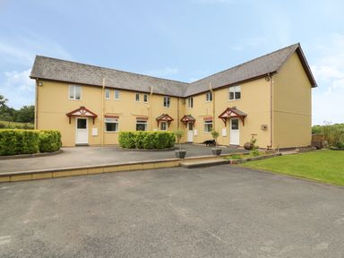 A two-story yellow building with multiple entrances and a paved driveway with shrubs at Foxes Meadow in Howey near Llandrindod Wells