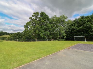 A grassy field with a soccer goal post and a paved area surrounded by trees at Foxes Meadow in Howey near Llandrindod Wells