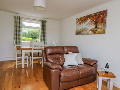 A brown leather sofa with pillows a small table with wine bottle and glasses a dining table with chairs and a window with curtains in a room at Foxes Meadow in Howey near Llandrindod Wells
