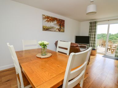A dining area with wooden table and white chairs a vase with flowers on the table a living area with brown chairs a TV and sliding glass doors leading to a patio at Foxes Meadow in Howey near Llandrindod Wells