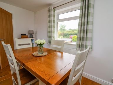 A dining room with a wooden table and white chairs a vase with flowers on the table and green check curtains at Foxes Meadow in Howey near Llandrindod Wells