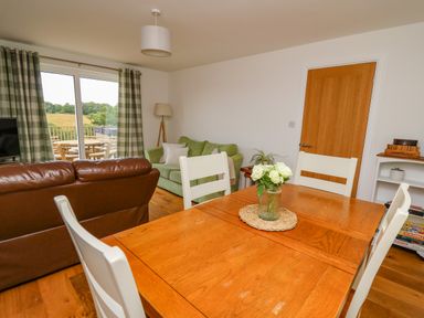 A dining area with a wooden table and white chairs a living room with green and brown sofas and a sliding glass door with checkered curtains at Foxes Meadow in Howey near Llandrindod Wells