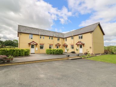 A beige two-story building with multiple entrances and small covered porches surrounded by shrubbery and a paved area at Pheasant Wood in Howey near Llandrindod Wells