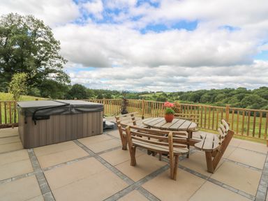 An outdoor patio with a wooden round table and benches a covered hot tub and a wooden railing overlooking a green landscape at Pheasant Wood in Howey near Llandrindod Wells