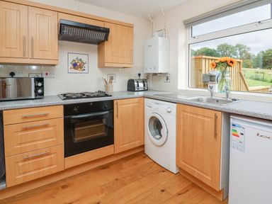 A kitchen with wooden cabinets a black oven a washing machine a toaster a microwave and a vase with flowers on the counter at Pheasant Wood in Howey near Llandrindod Wells