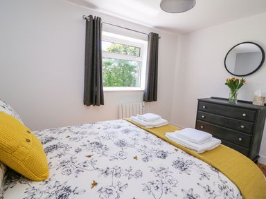 A bedroom with a bed with floral bedding and a yellow pillow beside a window with dark curtains and a black dresser with a round mirror and flowers at Owl Cottage in Llandrindod Wells