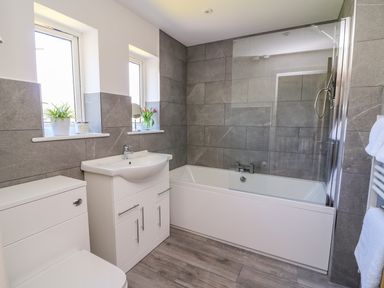 A bathroom with a toilet sink cabinet bathtub with glass panel and two windows with plants at Owl Cottage in Llandrindod Wells