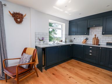 A kitchen with dark cabinets white tiled backsplash a window a wooden floor and a wooden chair with a cushion at Owl Cottage in Llandrindod Wells