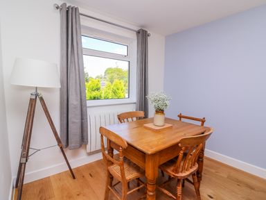 A dining area with a wooden table and four chairs a large window with gray curtains and a tripod floor lamp at Owl Cottage in Llandrindod Wells