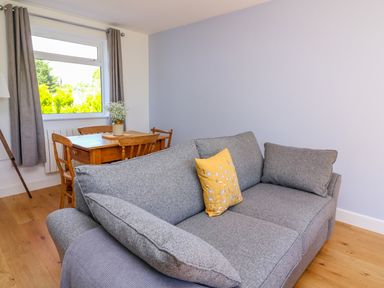 A gray sofa with a yellow pillow in a room with a wooden dining table and chairs near a window with gray curtains at Owl Cottage in Llandrindod Wells