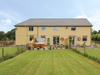 A backyard with green grass a hot tub two picnic tables and a yellow two story house at Owl Cottage in Llandrindod Wells