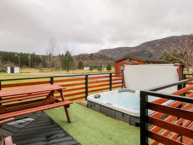 An outdoor deck with a wooden picnic table and a hot tub overlooking a grassy field and hills at Wildcat Lodge in Newtonmore