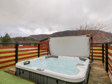An outdoor hot tub on a wooden deck with a partially open cover surrounded by a wooden fence and mountain view at Wildcat Lodge in Newtonmore