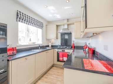 A kitchen with beige cabinets black countertops red appliances and a window with checkered curtain at Lilipad Cottage The Bay near Filey
