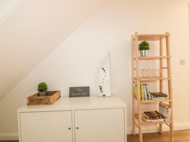 A white cabinet with a wooden tray labeled seashells and a small plant a sign that says The Beach Halt and a decorative piece next to a wooden shelving unit with books a plant and a basket at The Beach Halt in Perranporth